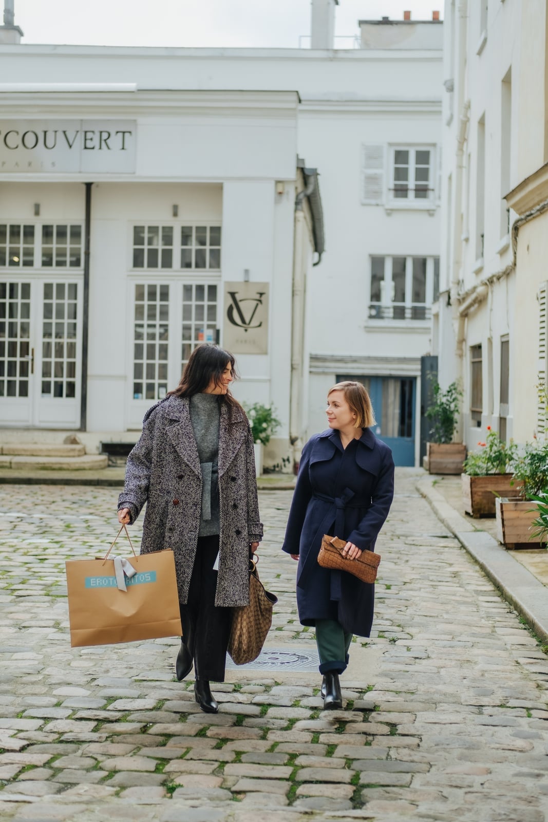 Two women carrying luxury shopping bags on cobblestone Paris street
